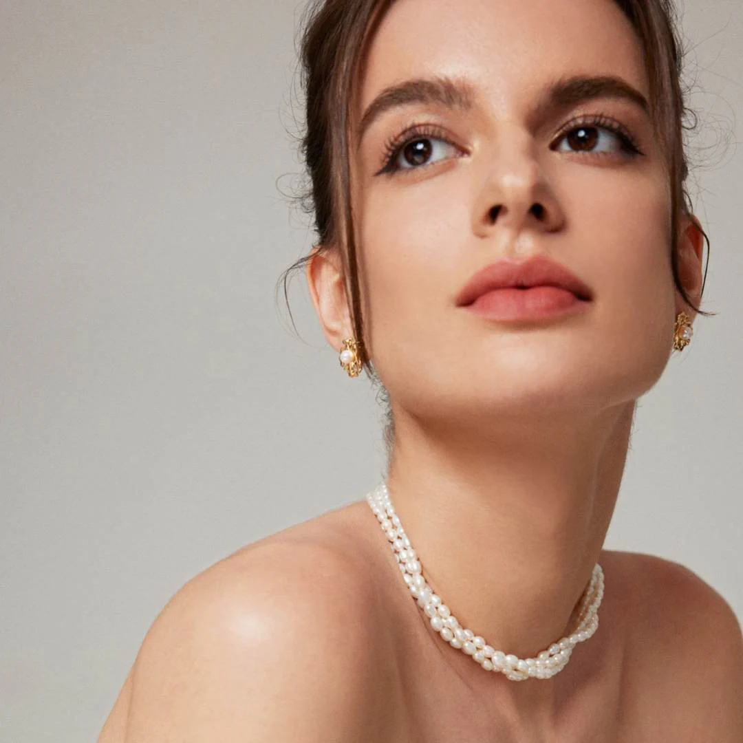 A woman in a flowing white dress, complemented by a triple-layer rice pearl necklace, radiating grace and charm.