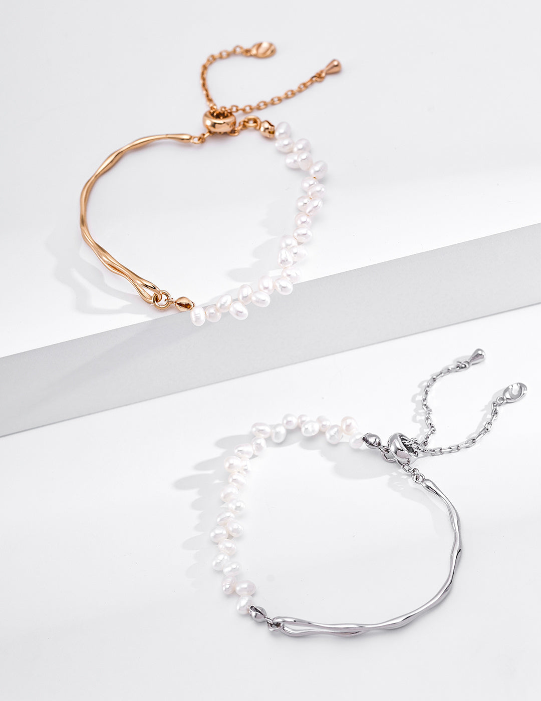 A clean studio shot displaying both the gold vermeil and silver variants of the Emily Bracelet side by side on a white surface, showcasing their asymmetric pearl and molten metal designs.