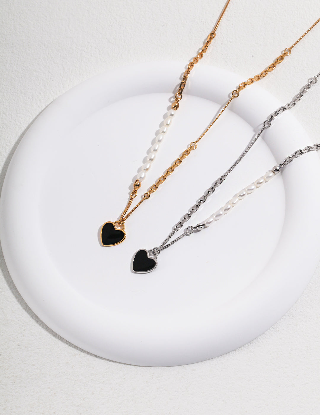 A white plate showcasing two necklaces with black heart charms - one gold, one silver - adorned with freshwater pearls.