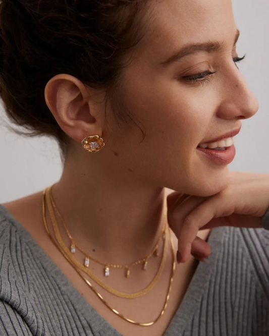 A woman with her hands on her face, adorned with a gold necklace featuring a cubic zirconia blooming flower design.