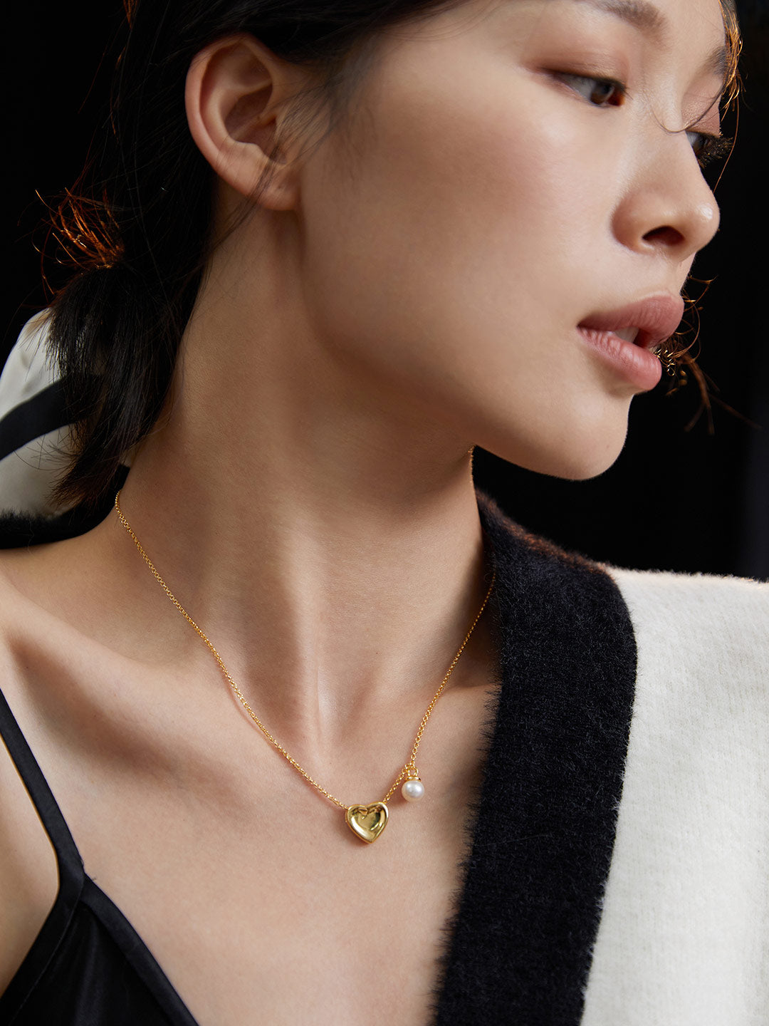 An elegant lady in a black sweater showcasing a gold necklace adorned with a heart pendant and freshwater pearl.