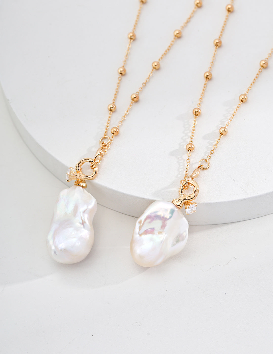 Studio product photograph showcasing a side-by-side comparison of two Genevieve gold necklaces, detailing the unique, organic variations in shape and luster of the large baroque pearl pendants.