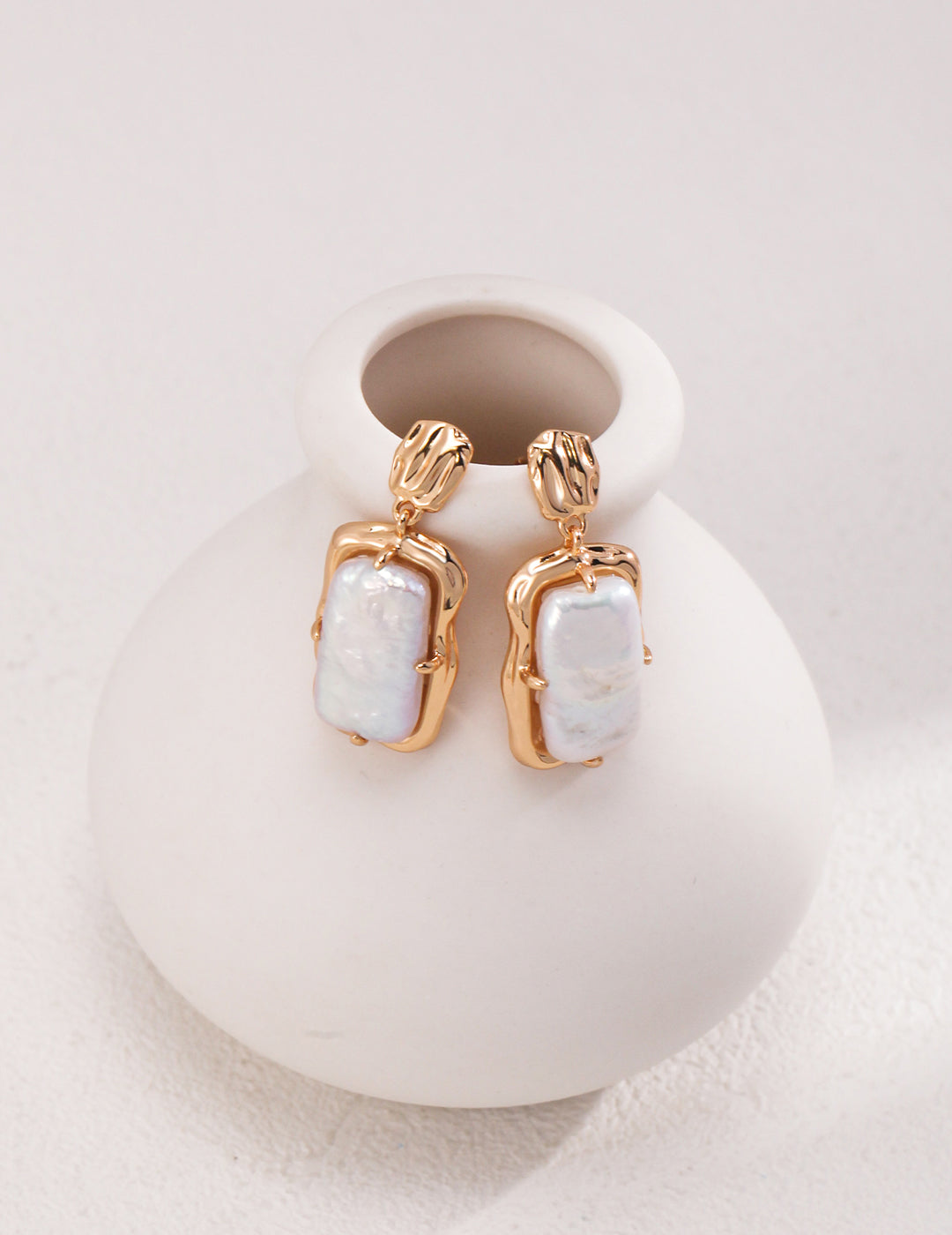 Gold plated pearl earrings elegantly placed on a white vase. Perfect blend of sophistication and beauty.