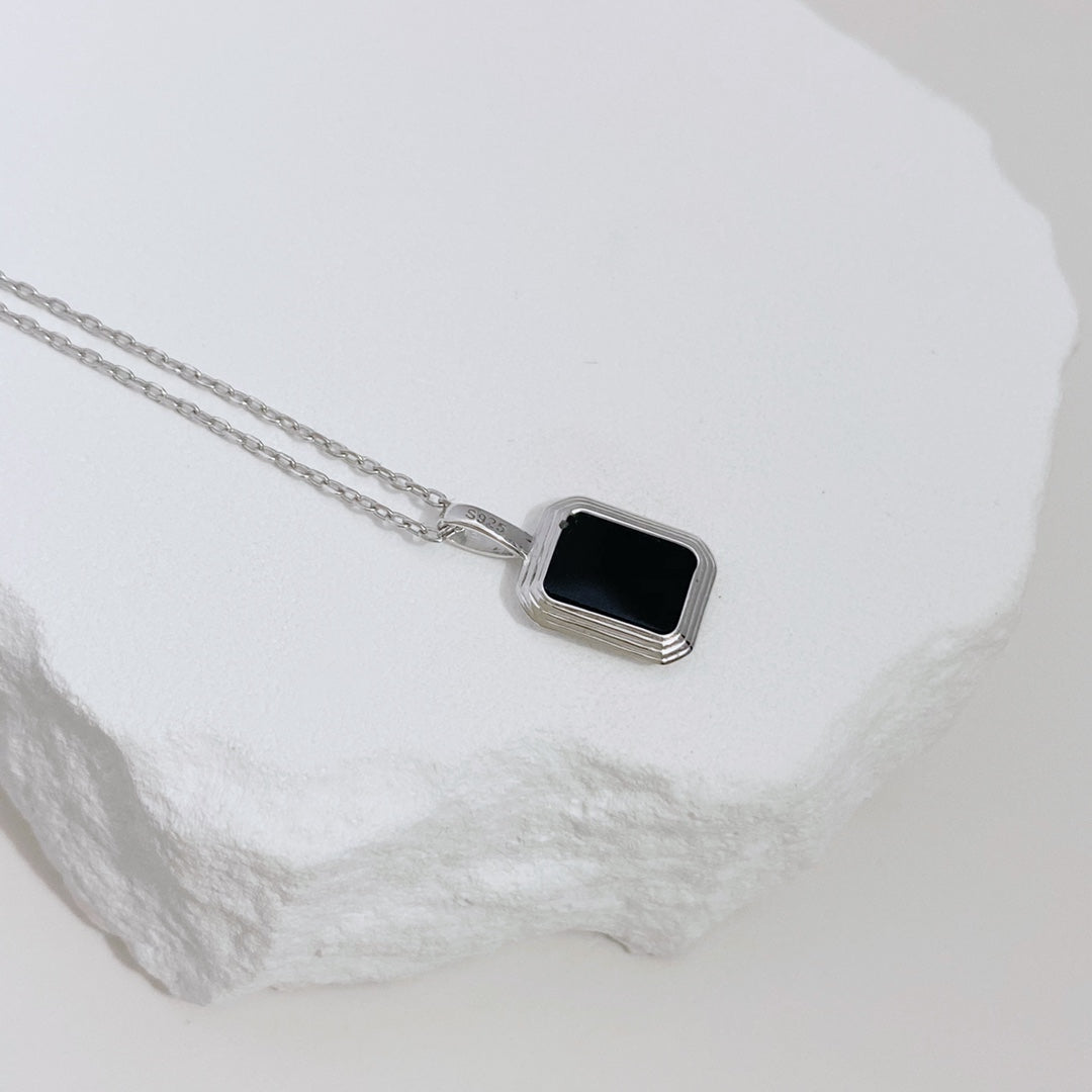 A silver necklace lies on a white rock, highlighting a reversible pendant of black agate and shell pearl.