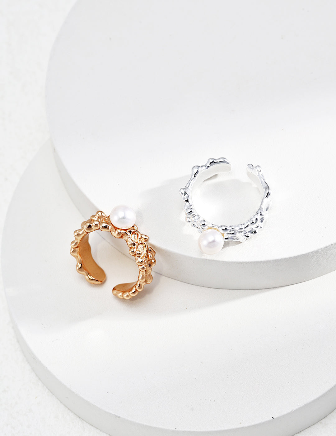 Flat lay studio view of two open-style Harper rings arranged on minimalist white geometric steps, showcasing the design in both textured gold and silver finishes, each topped with a single white pearl.