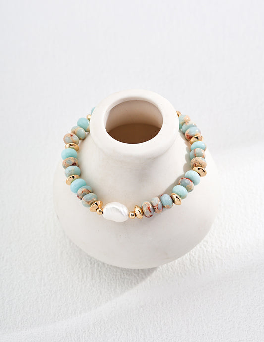 A gold plated pastel bracelet featuring blue agalmatolite stones and freshwater pearl hanging over a white vase.