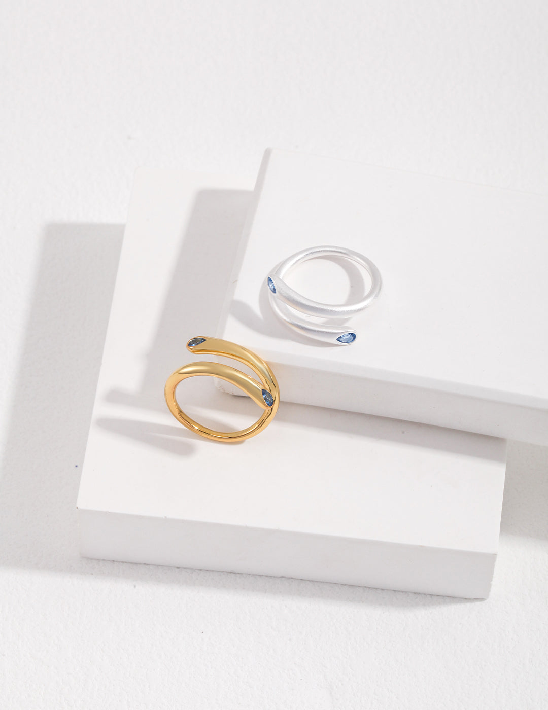 Two minimalist silver and gold wrap around rings with aquamarine stones on a white surface.