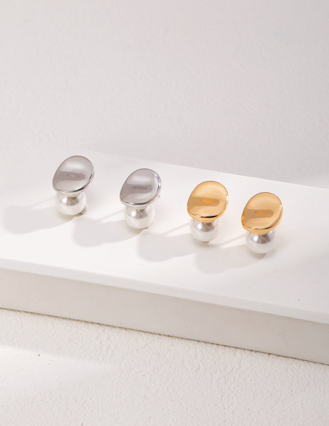 Pairs of jellybean studs with shell pearls on white surface, in silver and gold.