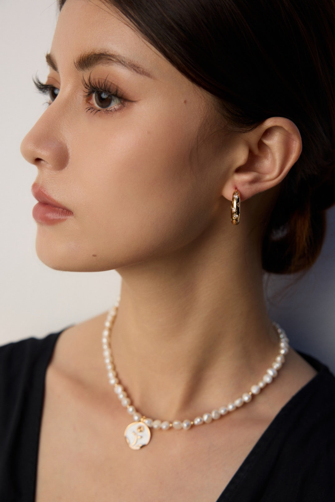 Lifestyle photograph of a model in a black top from a quarter-profile angle, showcasing the gold Jocelyn hoop earring paired with a delicate, uniform freshwater pearl pendant necklace.