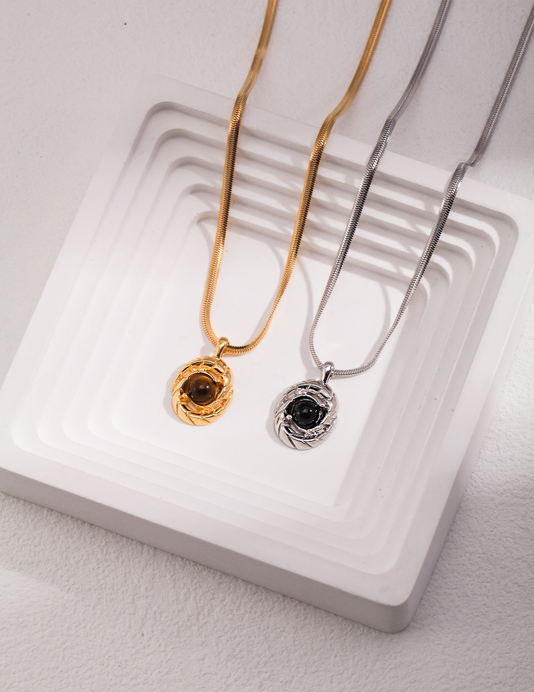 Two necklaces on white tray: gold with tiger's eye, silver with black agate stones.