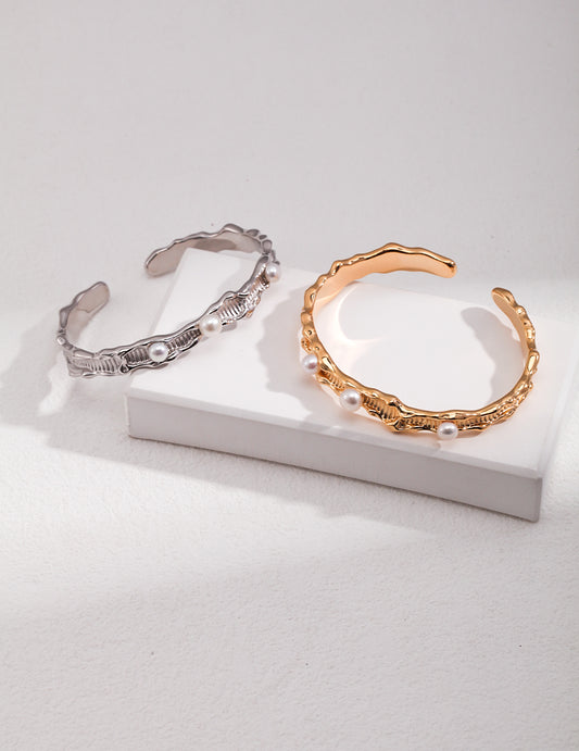 Two elegant cuff bracelets, one gold and one silver, resting on a white surface, both adorned with freshwater pearls.