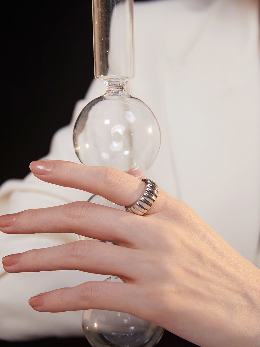 Female in white suit with glass vase, accessorized with silver gear wheel ring.