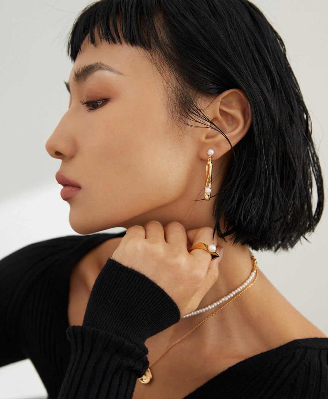 A stylish woman in a black sweater and gold earrings, wearing a square-shaped ring with a freshwater pearl and matching jewelry.