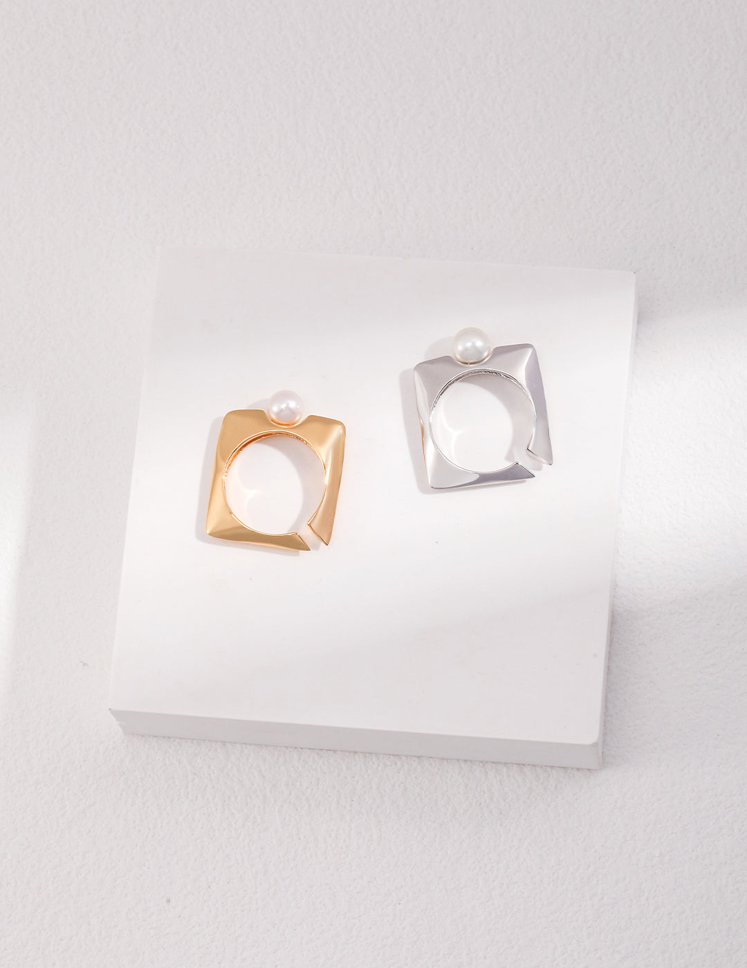 Two square-shaped rings with freshwater pearls, one silver and one gold, resting on a white surface.