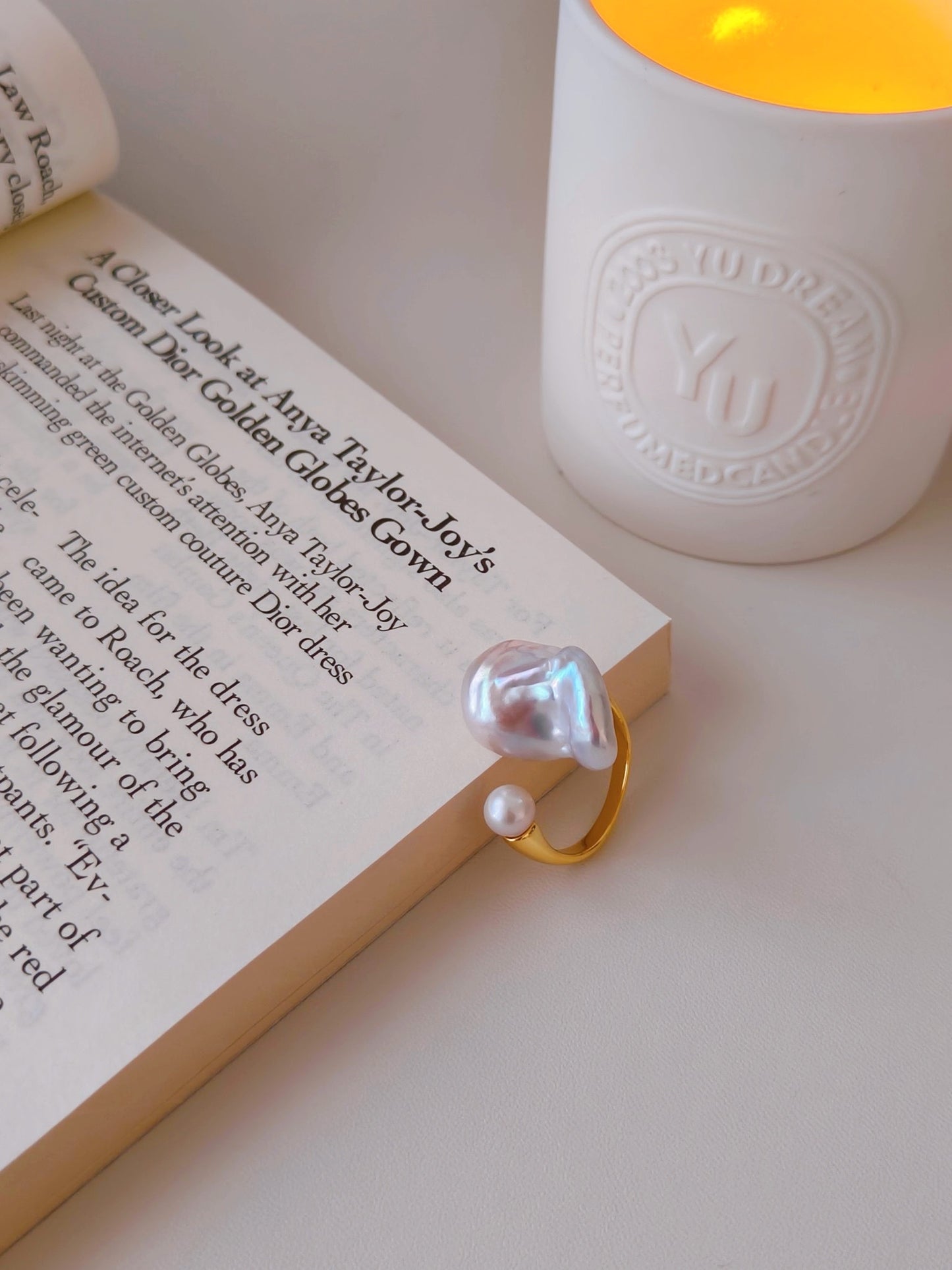 A baroque pearl ring rests on an open book beside a softly glowing candle, creating a serene and elegant atmosphere.