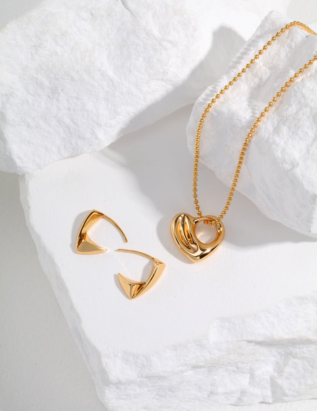 Gold jewelry set including hoop earrings and heart pendant necklace on white background.