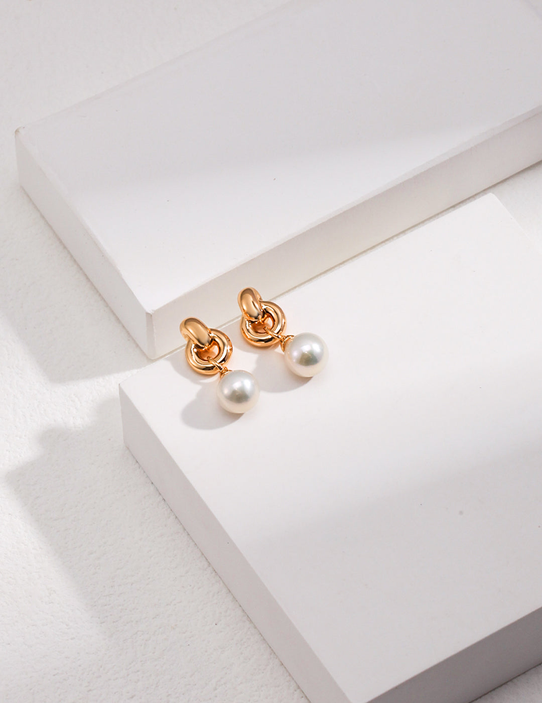 Two gold tone earrings with pearls on top, featuring gold interlocked knot design and freshwater pearl accents.