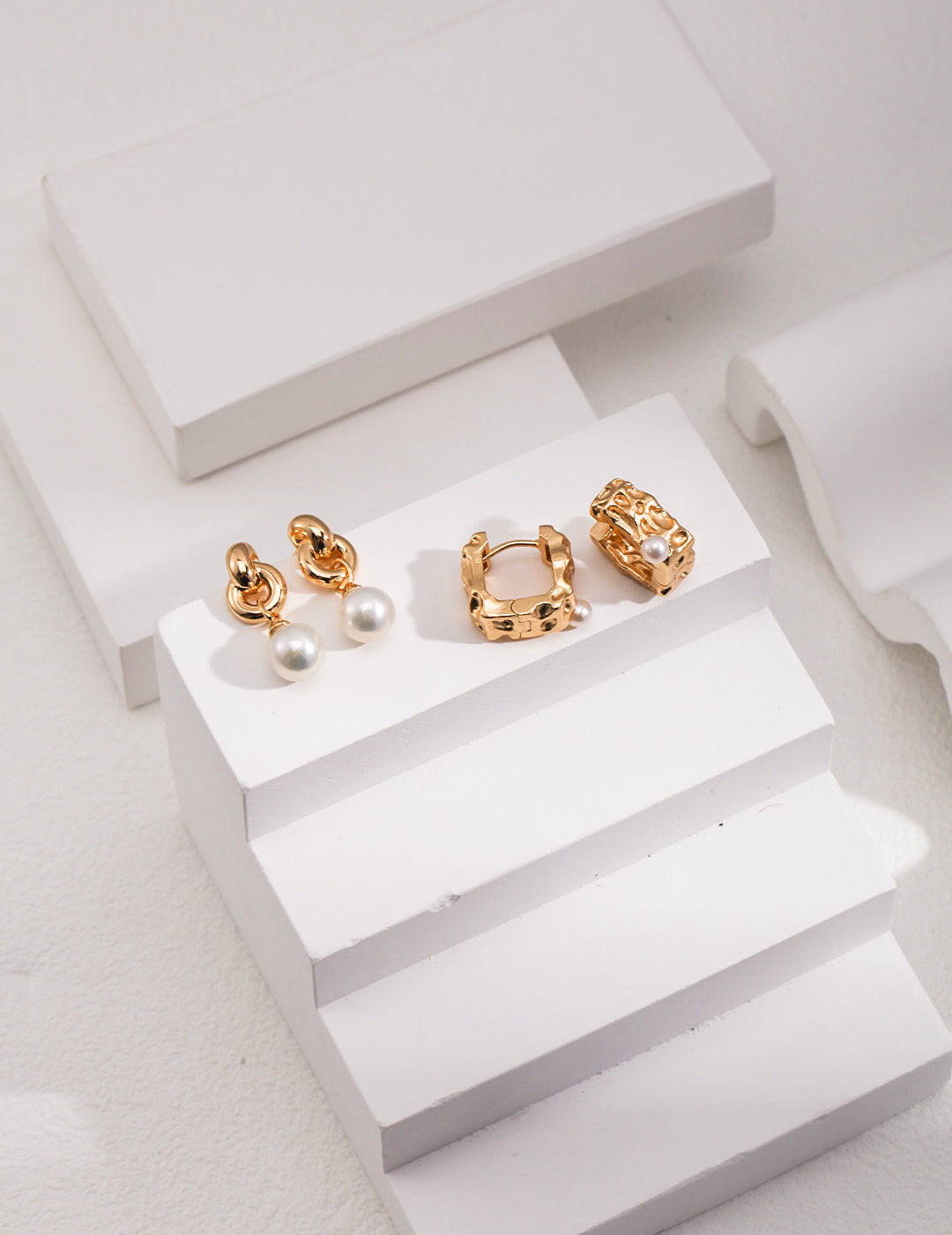 Two pairs of gold plated pearl earrings resting on white surface.