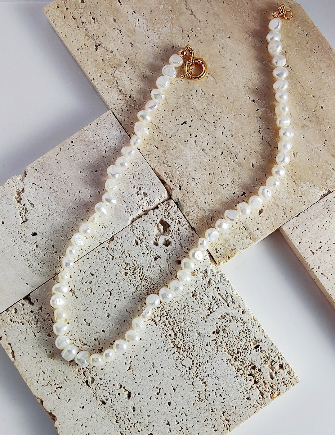 Miley Necklace featuring a strand of luminous freshwater baroque pearls with a gold circular clasp, laid out on textured light-colored stone tiles.