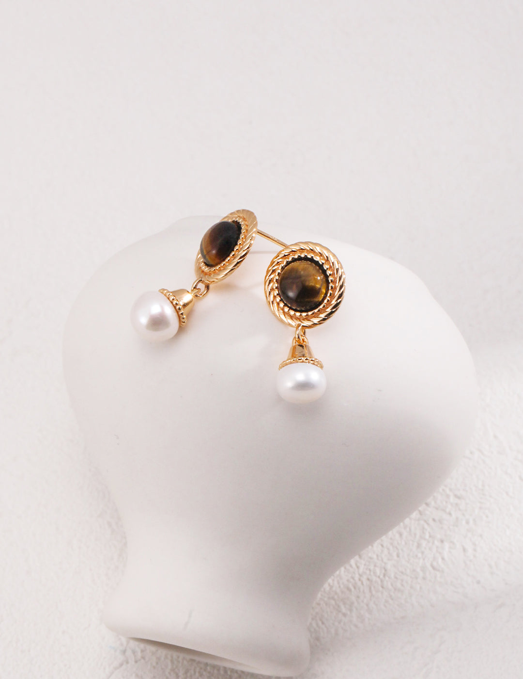 Gold plated round earrings with tiger's eye and freshwater pearls drop on a white vase.