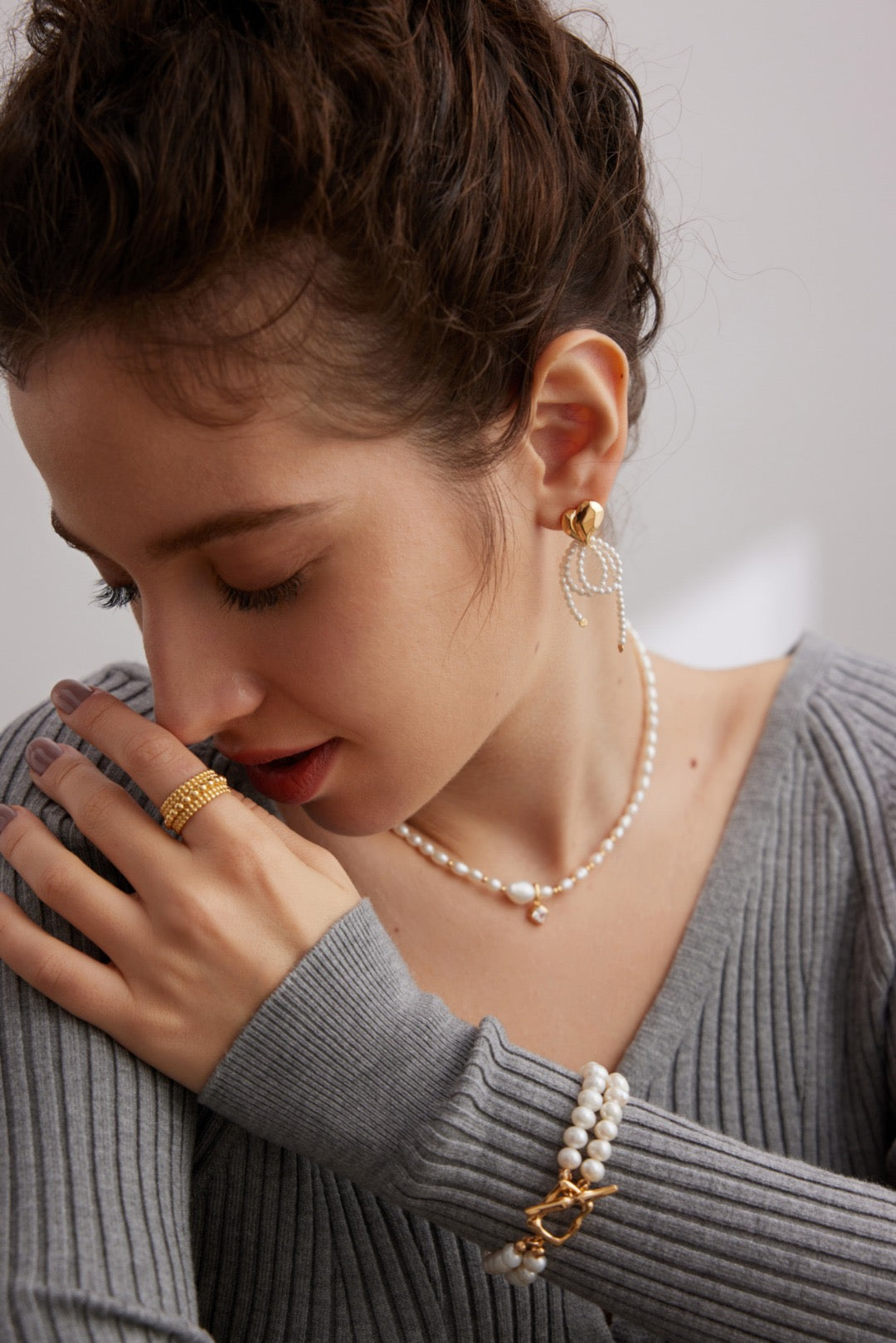 Woman wearing gold rings, earrings, and a pearl necklace, all from Mezuille, with a gray sweater.