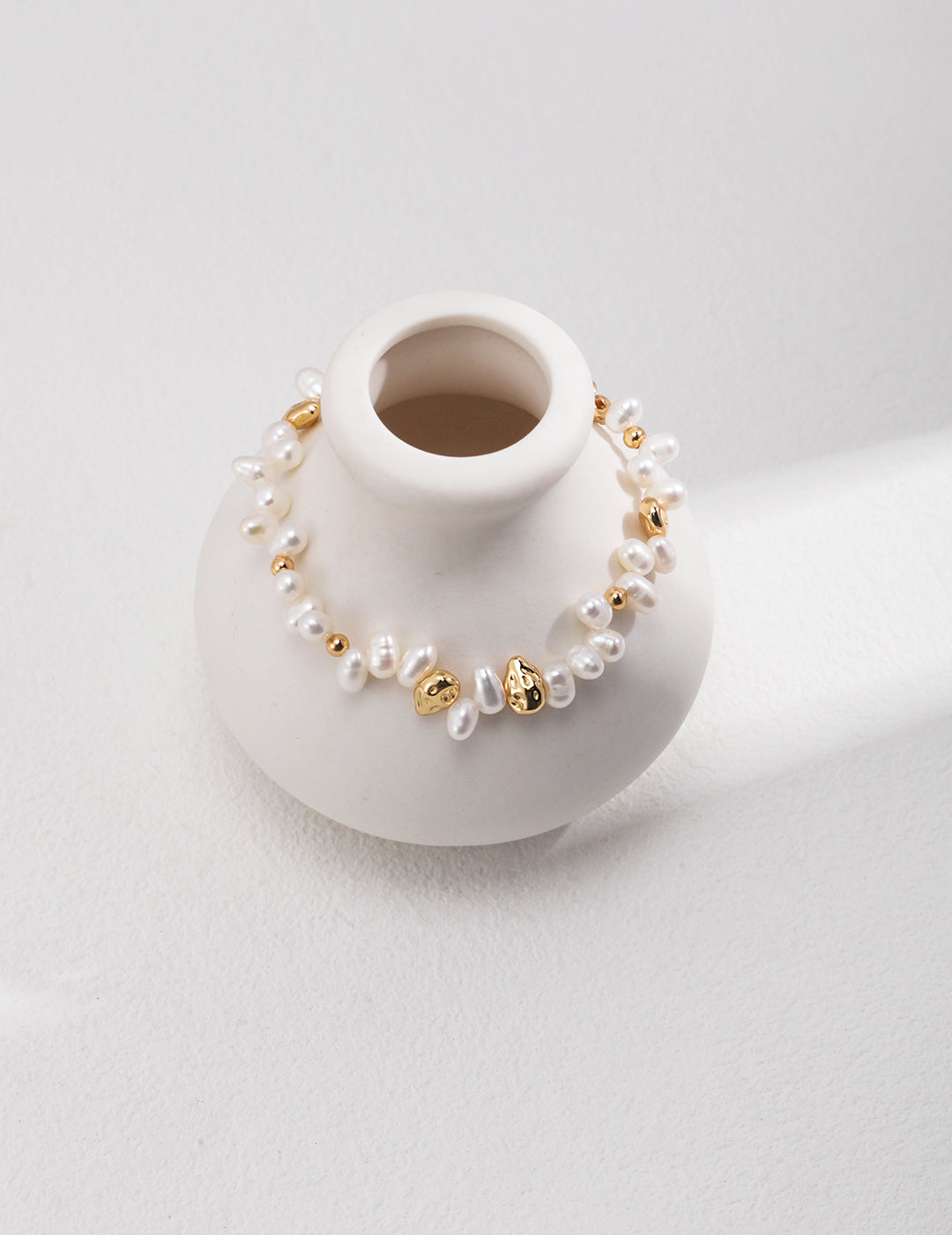 A bracelet adorned with gold beads and pearls, complemented by a white vase.