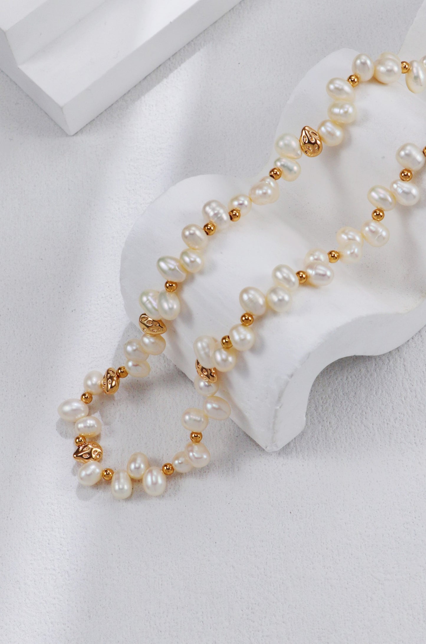 Studio product photograph of the Natasha pearl necklace casually draped over a white, wavy display riser, highlighting the repeating pattern of lustrous white pearls and gold spacers.