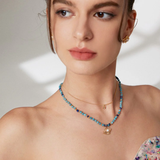 A woman adorned with a blue necklace, layered with dainty zirconia, and gold earrings, highlighting her chic fashion sense.
