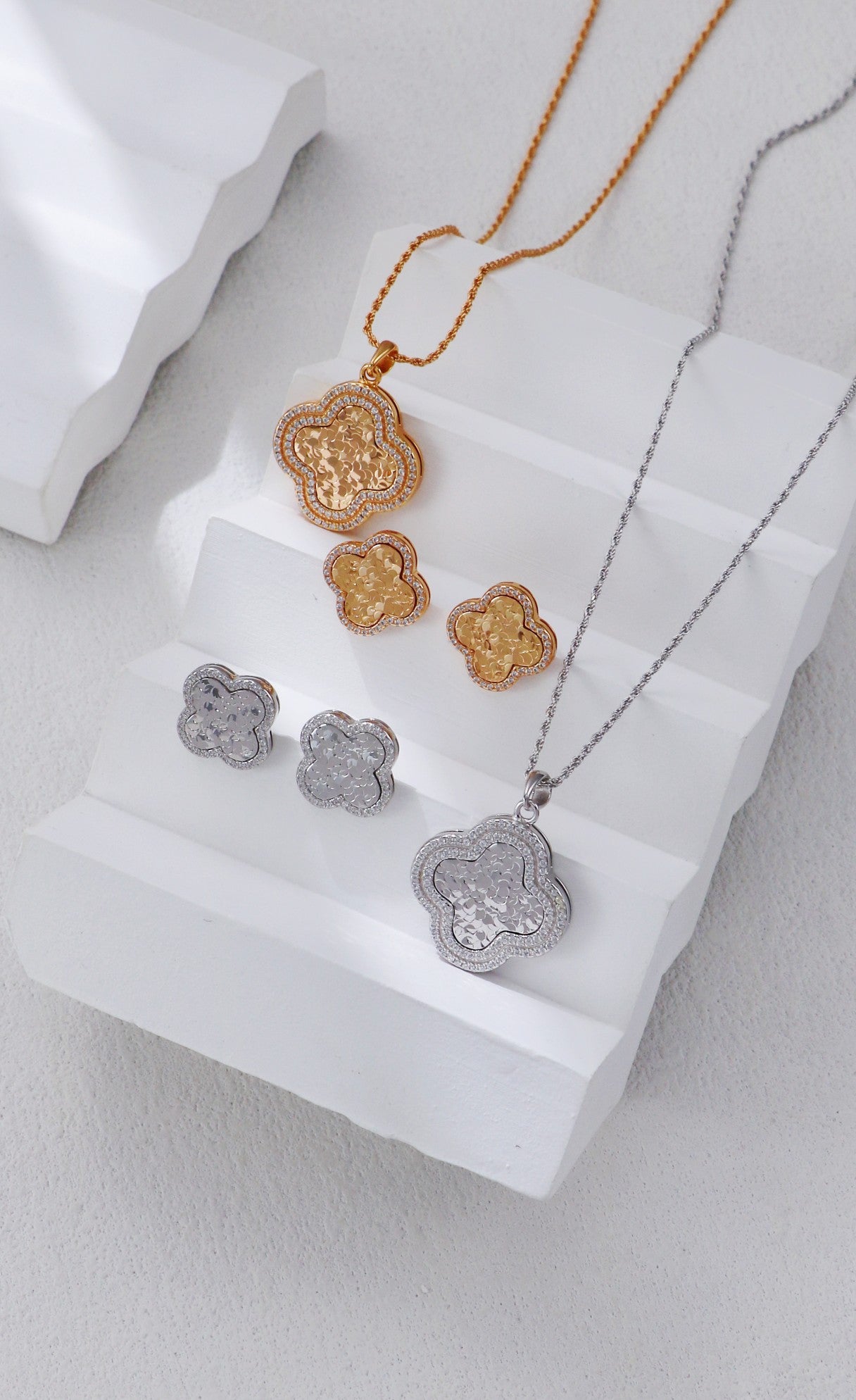 Two elegantly arranged clover-themed jewelry sets, including silver and gold necklaces and earrings, on a white surface.