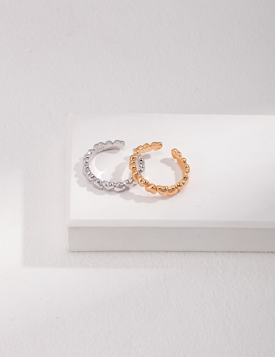 A silver and a gold ring, both with heart designs, resting on a white surface.