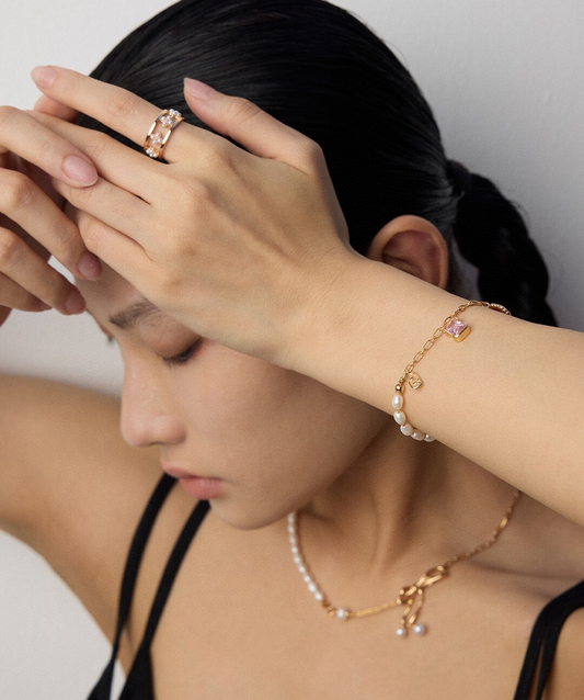 A model wearing Rebecca Bracelet—a gold bracelet with pearls and a pink stone—pairs it with a pearl necklace and a gold ring. Resting her hands on her forehead, she showcases elegant pieces perfect for everyday wear.
