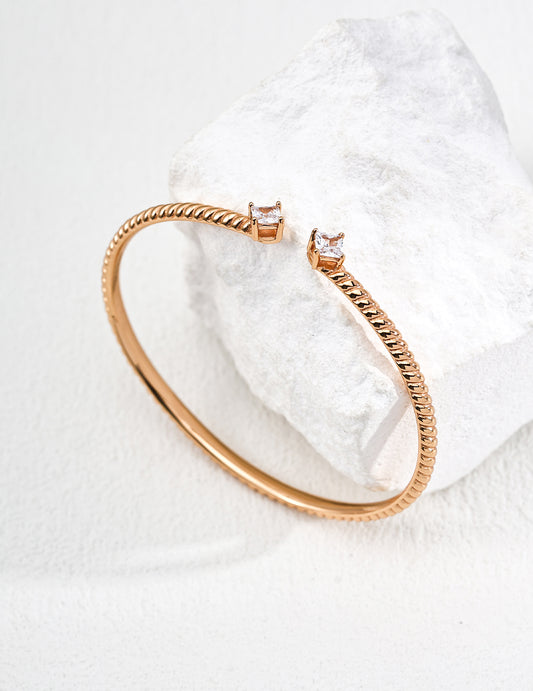A gold bangle with a textured band and two square-cut zirconia, elegantly wrapped around a rough white stone, set against a soft, neutral background.