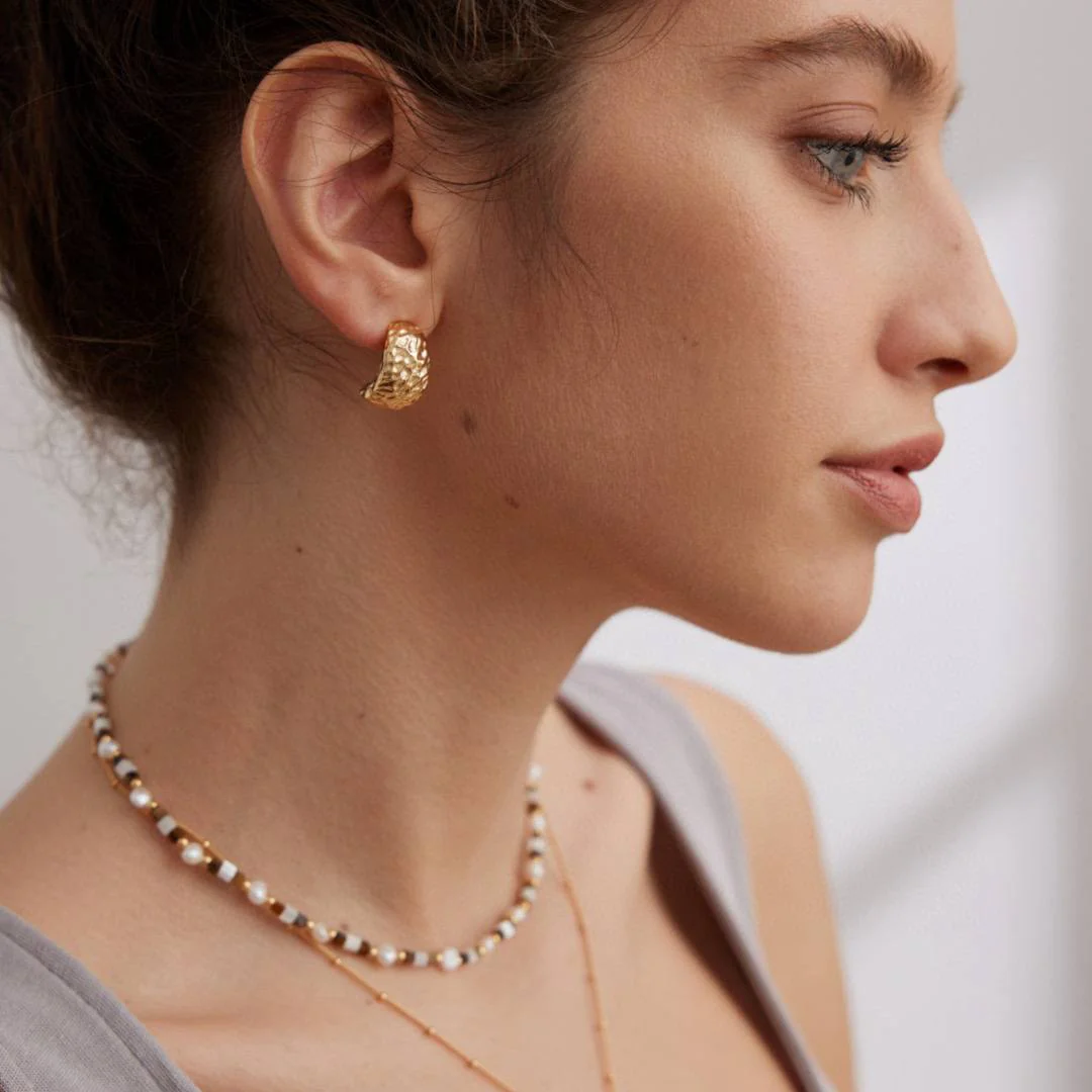 A woman displays a gold necklace and textured earrings, emphasizing her chic jewelry choice and personal style.