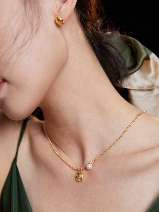 A woman wearing a gold necklace and earrings, with a gold chain necklace featuring a little bean pendant.
