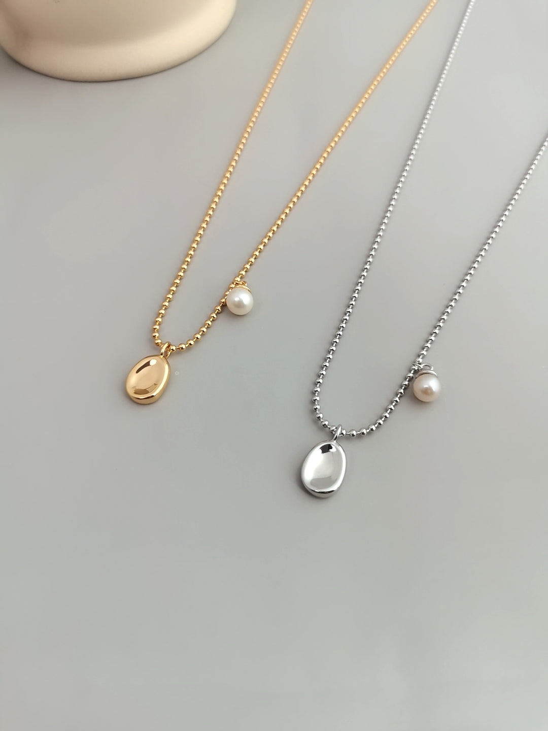 A pair of gold and silver necklaces adorned with dainty pearls and complemented by delicate bean pendants.
