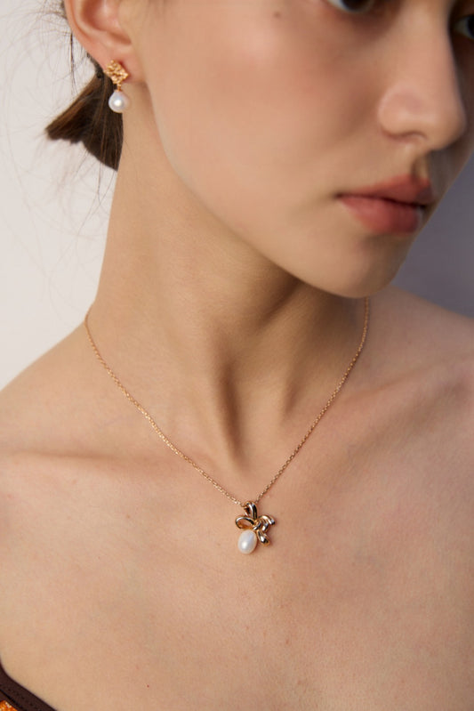 A woman showcases a gold necklace with a playful twist knot, adorned with a flower and pearl.