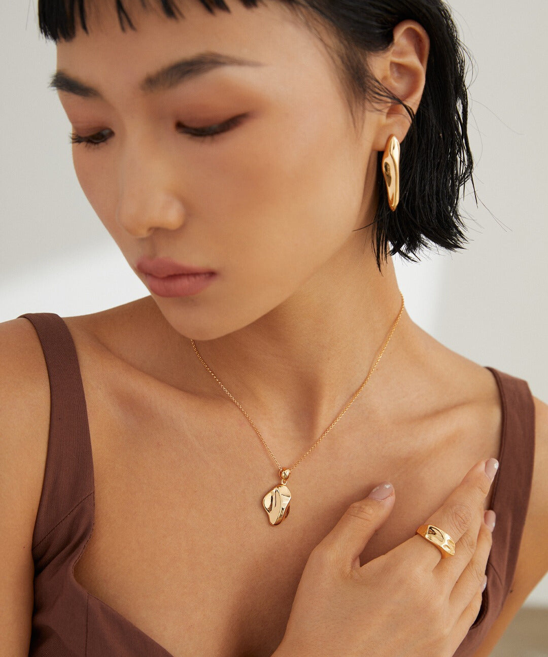 A woman wearing a gold necklace and earrings, with a model wearing a gold wavy design ring.