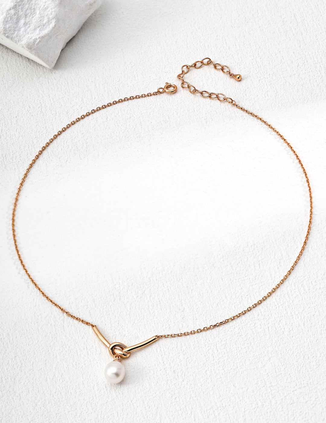 A product photograph of the 18K gold-plated Love Me Knot necklace, showcasing its delicate chain, sculptural knot accent, and dangling freshwater pearl pendant.   