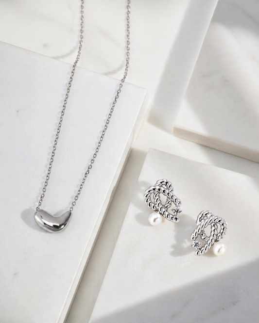 A styled product photograph of The Silver Duo set, featuring the Beanie heart pendant necklace and the Amélie pearl and rope stud earrings arranged together on neutral surfaces.