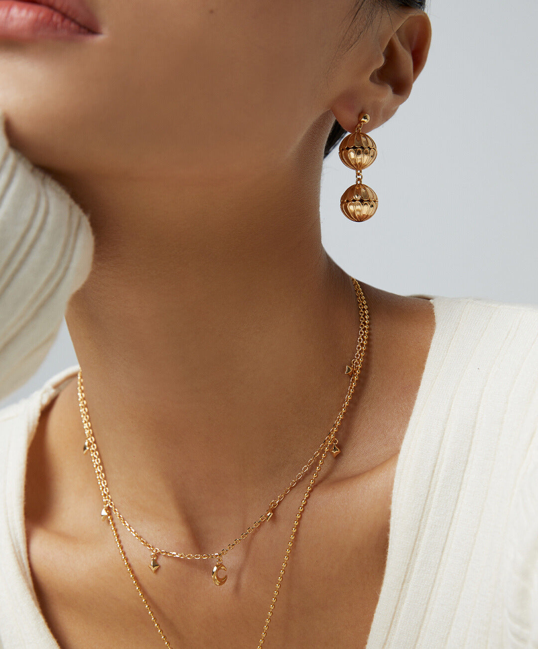 A woman in a white sweater and gold earrings, accessorized with a gold plated necklace featuring celestial charms.