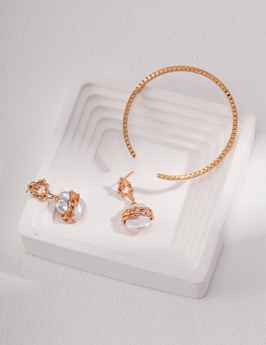 A white tray holding earrings with lustrous freshwater pearls and a gold bangle.