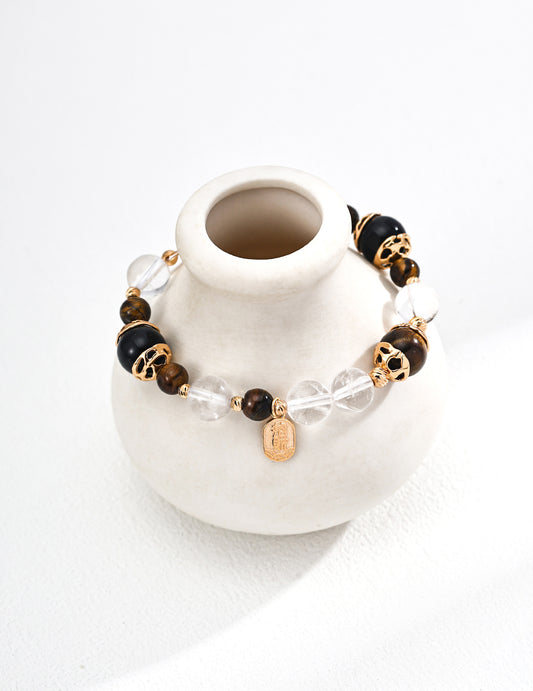 A stylish bracelet made of black and brown beads, clear crystals, gold accents, and a small pendant, resting on a white ceramic vase.