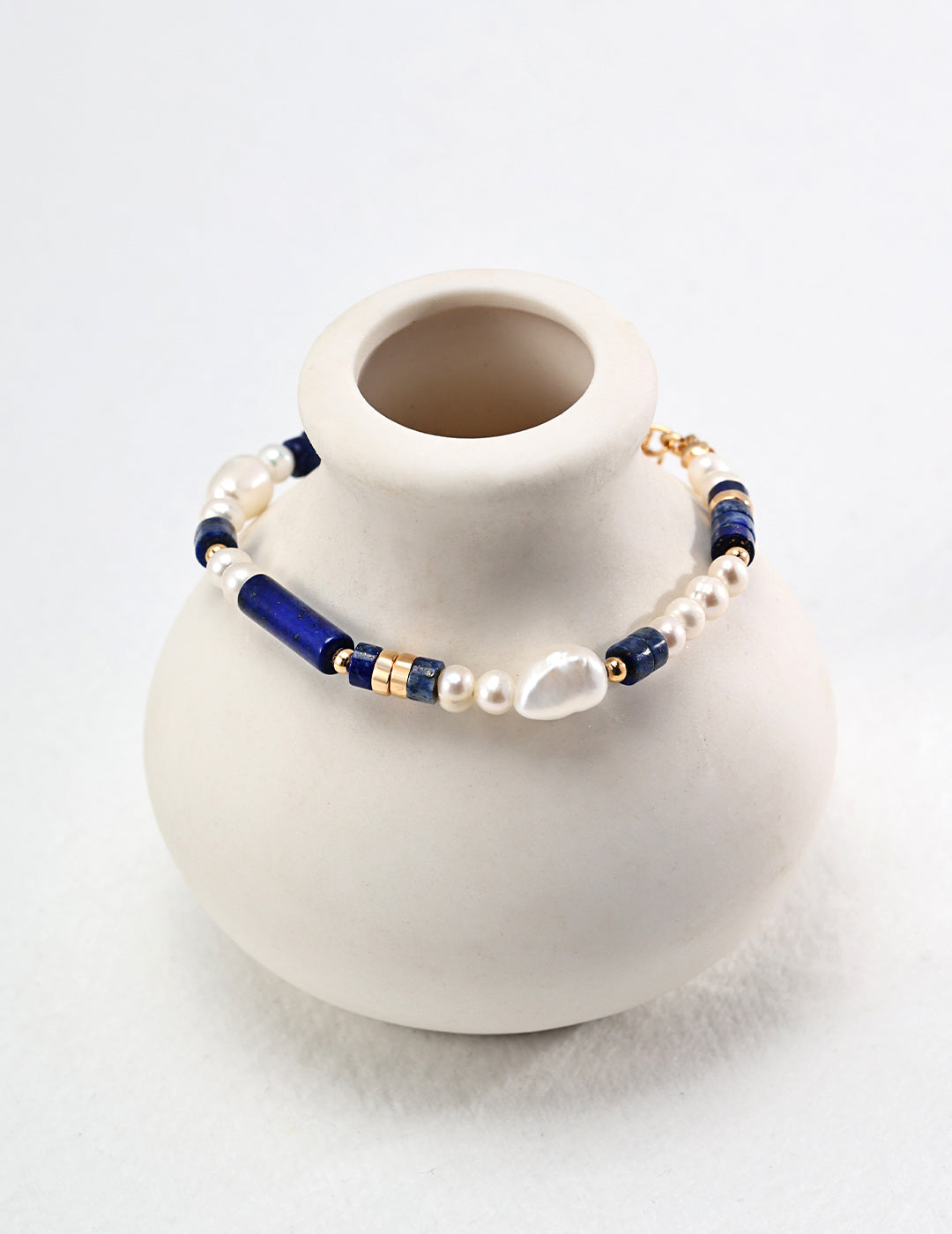 White vase holding blue and white bracelet beside gold plated bracelet adorned with freshwater pearls and lapis lazuli.