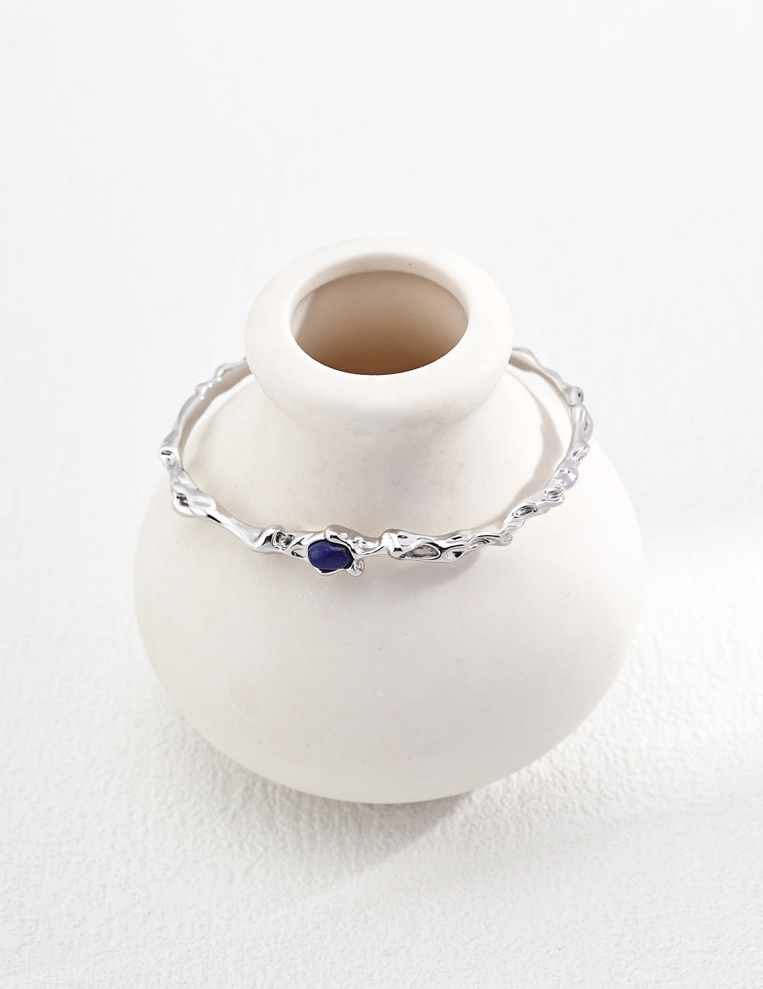 A sleek white vase holds a vibrant silver bangle featuring lapis lazuli, adding a touch of color to a clean, modern aesthetic.