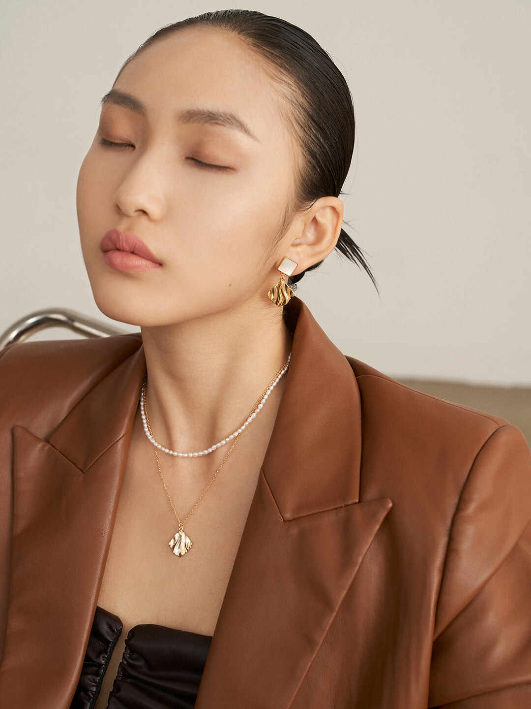 A stylish woman in a brown jacket and necklace, complemented by gold plated nacre earrings.