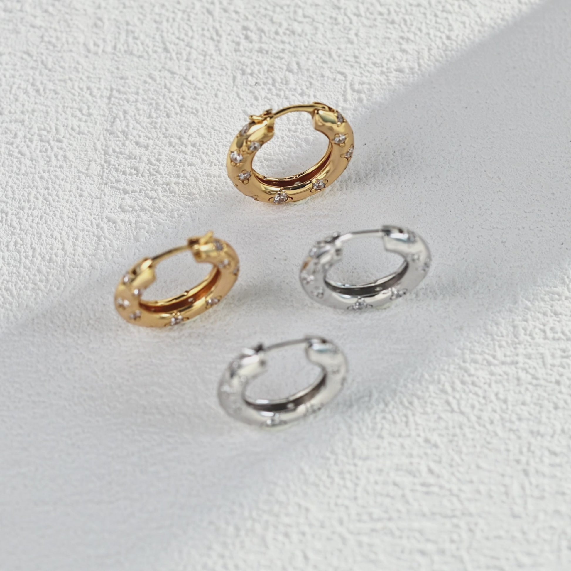 Close-up studio video showcasing the Jocelyn hoop earrings in both gold and silver finishes, rotating slowly on a textured white surface to highlight the sparkling star-set crystals and polished metal architecture.